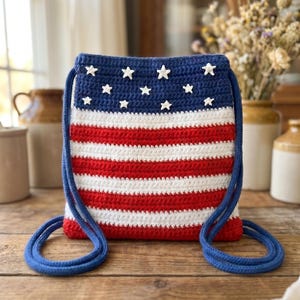 Puede incluir: Un bolso de ganchillo rectangular con cord&oacute;n que presenta un patr&oacute;n de rayas rojas, blancas y azules. La secci&oacute;n superior es azul s&oacute;lido con peque&ntilde;as estrellas de ganchillo blancas, mientras que la parte inferior tiene rayas horizontales.