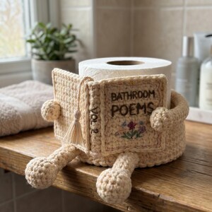 May include: A beige crochet toilet paper holder shaped like a book titled Bathroom Poems Vol 2. The cover features embroidered flowers and a small tassel. The holder has two arms and two legs extending from the base, designed to wrap around a roll.