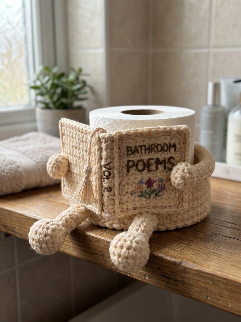May include: A beige crochet toilet paper holder shaped like a book titled Bathroom Poems Vol 2. The cover features embroidered flowers and a small tassel. The holder has two arms and two legs extending from the base, designed to wrap around a roll.