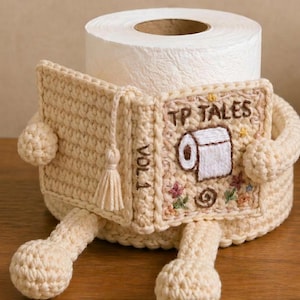 May include: A cream-colored crochet toilet paper roll cover shaped like a book with the title TP TALES VOL. 1 embroidered on the front. The cover features a small tassel, embroidered flowers, and a toilet paper roll graphic, with two dangling legs.