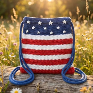 Puede incluir: Una mochila de crochet con cord&oacute;n que presenta un patr&oacute;n de rayas rojas, blancas y azules. La secci&oacute;n superior es azul s&oacute;lido con peque&ntilde;os adornos de estrellas blancas, mientras que la parte inferior consta de rayas horizontales rojas y blancas.