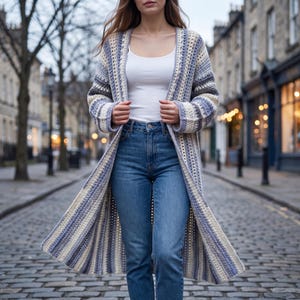 May include: A long, open-front crochet cardigan featuring vertical stripes in shades of blue, grey, and cream. The garment is worn over a plain white tank top and blue denim jeans, showcasing a textured, multi-colored yarn pattern throughout.