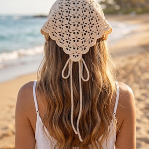 Può includere: Un foulard all'uncinetto color crema caratterizzato da un intricato motivo a ventaglio con bordo smerlato. Il copricapo triangolare include due lunghi lacci intrecciati che pendono lungo la schiena, consentendo una vestibilità regolabile.