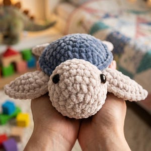 May include: A plush crochet sea turtle toy featuring a soft, textured beige head and flippers with a contrasting dark blue shell. The stuffed animal has two small, round, black plastic eyes set into the beige yarn, creating a simple, rounded form.