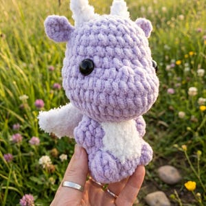 May include: A handmade, lavender crochet dragon with white accents, including wings and horns. The dragon has large black eyes and is held in front of a blurred background of green grass and wildflowers. A unique, whimsical plush toy.