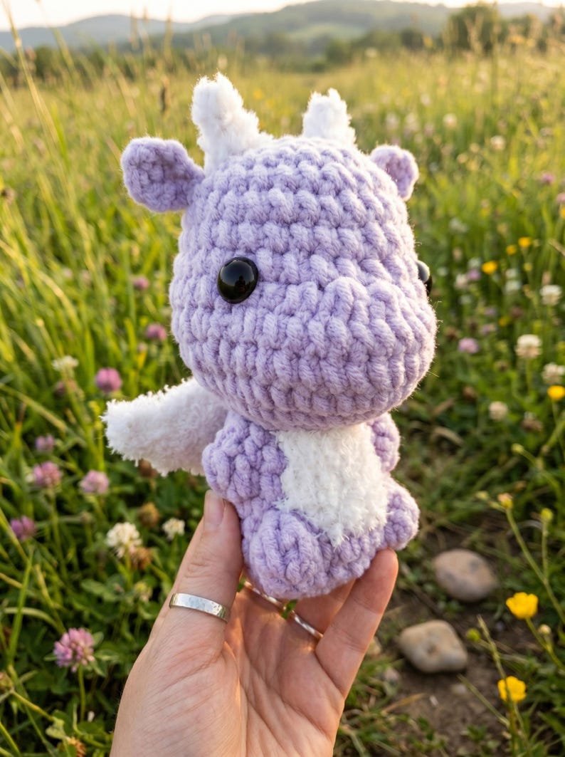 May include: A handmade, lavender crochet dragon with white accents, including wings and horns. The dragon has large black eyes and is held in front of a blurred background of green grass and wildflowers. A unique, whimsical plush toy.