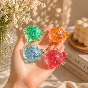 Peut inclure: Quatre figurines de tortues colorées remplies de paillettes, en vert, orange, bleu et rouge. Les tortues sont tenues dans une main, avec un vase de fleurs blanches et un livre intitulé "KINfolk ENTREPRENEUR" en arrière-plan.