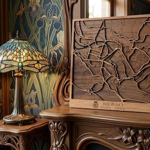 Peut inclure: Une carte en bois découpée au laser de Bilbao, Espagne, encadrée dans un cadre en bois. La carte est posée sur une surface en bois, à côté d'une lampe Tiffany en forme de libellule avec un abat-jour en vitrail. Le texte "BILBAO" est visible.