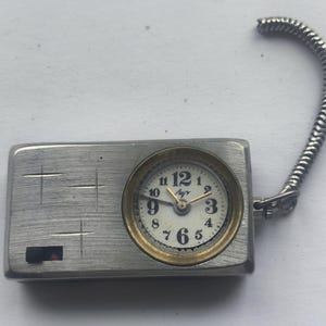 May include: A small, rectangular silver-toned watch with a round clock face. The clock face has gold-coloured trim, black numerals, and gold hands. A flexible, silver-toned metal loop is attached to the watch.
