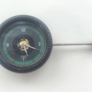 May include: A vintage black and green analog clock with a silver stem and a dark blue knob. The clock face has white numerals and green accents. The clock is round and has a textured black bezel.