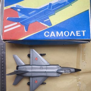 Puede incluir: Un avión de juguete gris con emblemas de estrella roja y una nariz negra se muestra junto a su caja. La caja es azul, amarilla y roja, con una ilustración de avión y la palabra "CAMOЛET".