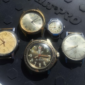 Puede incluir: Colección de cinco relojes de pulsera vintage. Los relojes presentan varios diseños de esfera, incluyendo números romanos y arábigos. Las esferas son de color dorado, plateado y negro, con correas de metal. Los relojes están dispuestos sobre una superficie oscura.