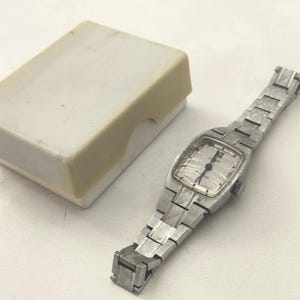 May include: A silver-toned analog wristwatch with a rectangular face and a textured metal link bracelet, accompanied by a small, rectangular, off-white plastic storage box. The watch dial features thin black hands and simple hour markers.