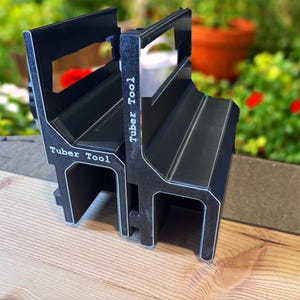 May include: Two black Tuber Tool devices featuring a rigid, angular frame with a hollow base and a vertical back panel. The text Tuber Tool is printed in white on the side of each unit. The devices have a matte finish with visible white edges.
