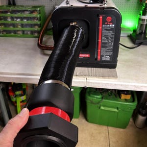 May include: A black and red diesel air heater with a digital display showing temperature. The heater has a black exhaust pipe and is connected to a black and red fitting. Tools and storage containers are visible in the background.