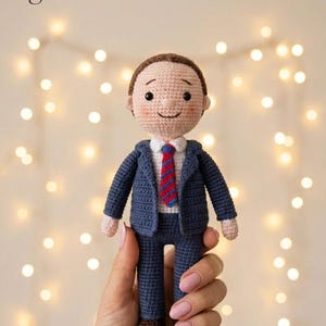 May include: A crochet doll featuring a person with brown hair, wearing a dark blue suit, a white shirt, and a red and blue striped tie. The doll has black bead eyes and pink cheeks. The text gullabstoreco is visible in the upper left corner.