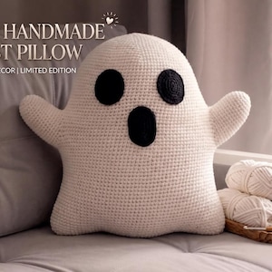 May include: A cream-colored crocheted ghost pillow with two round black eyes and an oval black mouth. The text COZY HANDMADE GHOST PILLOW AMIGURUMI DECOR | LIMITED EDITION is visible. Beside the pillow are two white yarn balls and one black yarn ball.