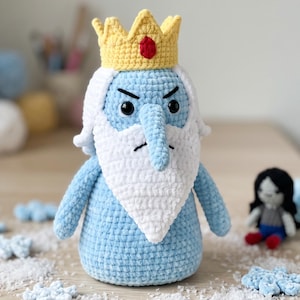 May include: A crocheted plush figure of a character with light blue skin, a long pointed nose, and a white beard. The figure wears a yellow crown with a red gem in the center. It has black embroidered eyes and eyebrows, and a small black mouth.