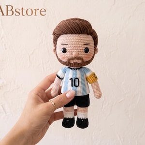 Puede incluir: Una muñeca de ganchillo hecha a mano que representa una figura masculina con cabello castaño y barba, vistiendo una camiseta de fútbol a rayas azul claro y blanca con el número 10, pantalones cortos negros y un brazalete de capitán amarillo.