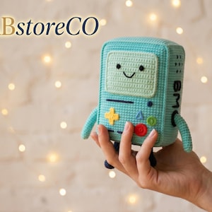 May include: A light blue crocheted BMO character toy with a rectangular body, a screen face with a stitched smile, and colorful buttons including a yellow cross, blue triangle, and red circle. The side features the text BMO in black stitching.