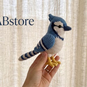 以下が含まれることがあります： 青、白、黒の糸で編まれたアオカケスの鳥のフィギュア。尖った冠羽、暗い色のくちばし、黄色い足が特徴です。画像の左上隅には、濃い青色のフォントでGullABstoreというテキストが表示されています。