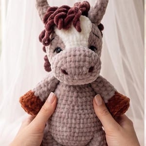 Puede incluir: Un juguete de caballo tejido a ganchillo con hilo suave de color marrón claro y una mancha blanca en la frente. Presenta hilo marrón oscuro en la crin, los cascos y la cola, con ojos de seguridad negros y nariz bordada en punto grueso.