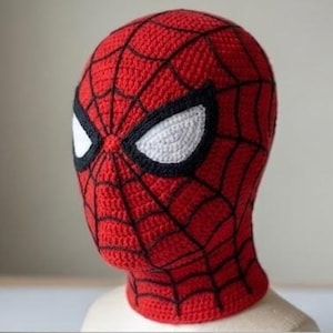 Pode incluir: Uma balaclava de crochê vermelha com um padrão de teia de aranha preta na superfície e dois grandes remendos oculares brancos em forma de amêndoa contornados com fio preto grosso, projetada para cobrir a cabeça e o pescoço com textura.