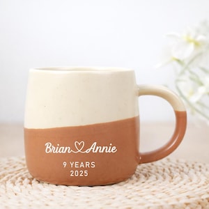May include: A two-tone ceramic mug with a cream upper section and a brown lower section. The mug is inscribed with "Brian & Annie" and a heart symbol, plus "9 YEARS 2025" in white. The handle is brown.