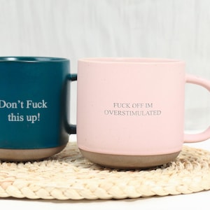 Puede incluir: Una taza de cerámica azul verdoso oscuro con el texto Don't Fuck this up! y una taza de cerámica rosa claro con el texto FUCK OFF IM OVERSTIMULATED. Ambas tazas tienen un acabado mate liso en la parte superior y una base marrón rugosa sin esmaltar.