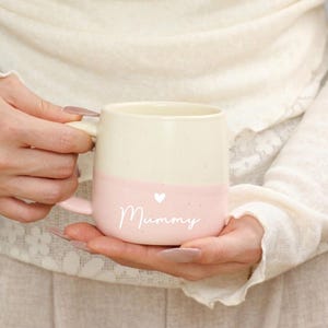 Puede incluir: Taza de cerámica de dos tonos, crema en la parte superior y rosa en la inferior. La palabra "Mummy" está escrita en cursiva en la sección rosa, con un pequeño corazón encima. La taza tiene un asa curvada.