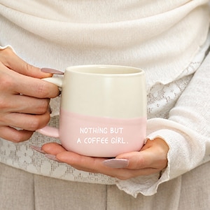 Könnte beinhalten: Ein zweifarbiger Keramikbecher mit einem cremefarbenen oberen Teil und einem hellrosa unteren Teil. Der Text "NOTHING BUT A COFFEE GIRL." ist in Weiß auf dem rosa Teil des Bechers aufgedruckt. Der Becher hat einen Henkel auf der linken Seite.