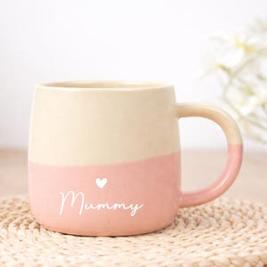 Op de afbeelding: Tweekleurige keramische mok met een crèmekleurige bovenkant en een roze onderkant. Het woord "Mummy" staat in witte sierletters geschreven, met een klein wit hartje erboven. Het handvat is ook roze.