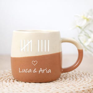 Könnte beinhalten: Eine zweifarbige Keramiktasse mit cremefarbenem Oberteil und braunem Unterteil. Die Tasse hat weiße Strichlisten, ein Herz und die Namen "Luca & Aria" in weißer Schrift. Der Griff ist braun.