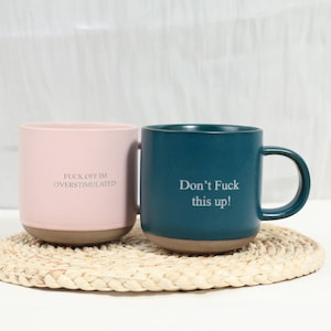 Op de afbeelding: Een lichtroze mok met de tekst FUCK OFF IM OVERSTIMULATED en een donkergroenblauwe mok met de tekst Don't Fuck this up!. Beide mokken hebben een gladde matte afwerking, een afgerond handvat en een contrasterende ongeglazuurde bruine onderkant.