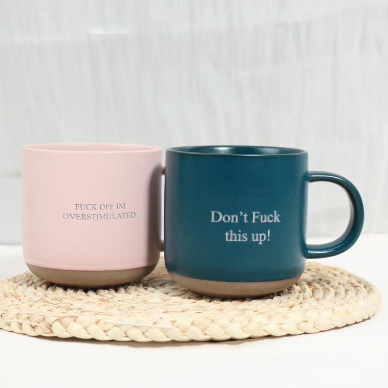 Op de afbeelding: Een lichtroze mok met de tekst FUCK OFF IM OVERSTIMULATED en een donkergroenblauwe mok met de tekst Don't Fuck this up!. Beide mokken hebben een gladde matte afwerking, een afgerond handvat en een contrasterende ongeglazuurde bruine onderkant.