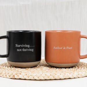 Op de afbeelding: Twee keramische mokken met oren, &eacute;&eacute;n zwarte en &eacute;&eacute;n terracotta oranje. De zwarte mok heeft de witte tekst "Surviving..... not thriving" en de oranje mok heeft de witte tekst "Author & Poet". Beide mokken hebben een natuurlijke beige basis.