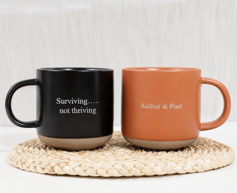 Op de afbeelding: Twee keramische mokken met oren, &eacute;&eacute;n zwarte en &eacute;&eacute;n terracotta oranje. De zwarte mok heeft de witte tekst "Surviving..... not thriving" en de oranje mok heeft de witte tekst "Author & Poet". Beide mokken hebben een natuurlijke beige basis.