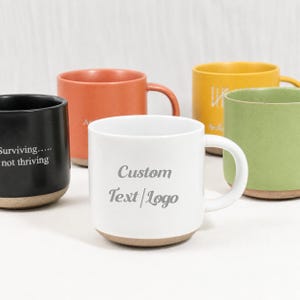 Op de afbeelding: Een verzameling van vijf keramische mokken in zwart, oranje, geel, groen en wit. De zwarte mok bevat de tekst 'Surviving... not thriving' en de witte mok toont de tekst 'Custom Text | Logo' in het grijs. Elke mok heeft een beige onderkant.
