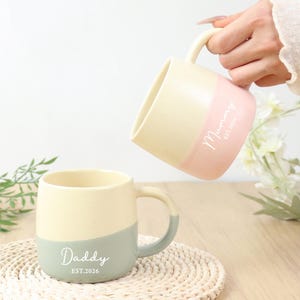 Op de afbeelding: Tweekleurige keramische mokken, waarvan er één wordt vastgehouden, met een crème- en roze kleurenschema. De andere mok is crème en grijs. De mokken hebben de woorden "Daddy EST.2026" en "Mummy EST.2026" erop gedrukt. De mokken staan op een geweven onderzetter.