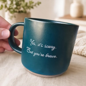 Op de afbeelding: Een donkergroenblauwe keramische mok met een matte afwerking en een gebogen handvat. De voorzijde van de mok bevat witte gegraveerde tekst die luidt: "Yes, it's scary. But you're brave." De basis heeft een lichte, ongeglazuurde keramische rand.