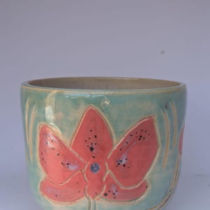 Puede incluir: Un cuenco de cerámica con un esmalte verde azulado claro y un gran diseño floral de color coral. La flor tiene motas negras y un centro azul. El borde del cuenco es de color beige neutro.