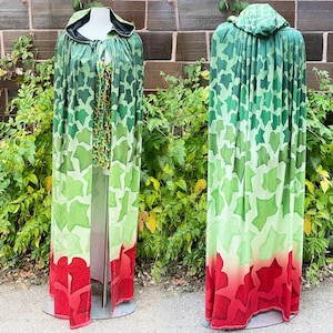 Pode incluir: Uma capa longa com capuz com um padrão de folhas verdes e vermelhas. A capa apresenta um design gradiente, que transita do verde para o vermelho na bainha. A capa é exibida em um manequim, mostrando seu design fluido.