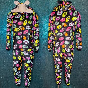 May include: A black one-piece jumpsuit featuring a colourful all-over pattern of stylised eyes in shades of pink, yellow, orange, and light blue. The garment includes a hood with small ears, a front zip closure, and long sleeves with cuffs.