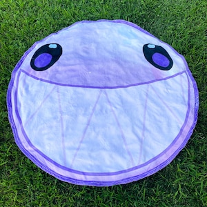 May include: A circular light purple blanket featuring a large, smiling face with two dark purple oval eyes and a wide mouth filled with triangular teeth. The fabric has a soft, plush texture and a distinct border line around the entire edge.
