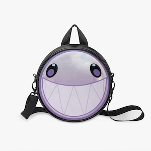 Può includere: Uno zaino rotondo nero caratterizzato da un volto circolare viola chiaro con un ampio sorriso dentato e grandi occhi viola scuro. La borsa include un manico superiore nero e spallacci neri regolabili fissati ai lati con ganci.