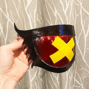 Könnte beinhalten: Eine schwarz-rote Maske mit einem gelben X-Design. Die Maske hat einen gezackten Rand und eine gebogene hornartige Verlängerung an der Seite. Die Maske wird von einer Hand gehalten.