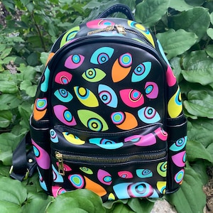 Pode incluir: Uma mochila preta com um padrão repetitivo de olhos estilizados em cores vibrantes, incluindo rosa neon, azul brilhante, verde-limão e laranja. A bolsa inclui um compartimento principal com zíper, um bolso frontal menor e bolsos laterais.