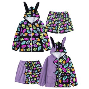 Pode incluir: Dois conjuntos de roupa compostos por casacos com fecho e orelhas de coelho no capuz e calções a condizer. As peças apresentam um fundo preto com um padrão colorido de olhos estilizados em tons de rosa, amarelo, azul e roxo.