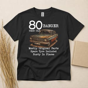 May include: Black t-shirt featuring a vintage car graphic with the text "80 BANGER YEAR OLD". The car is brown, orange, and blue. Text below reads "Mostly Original Parts, Spare Tyre Included, Rusty In Places".