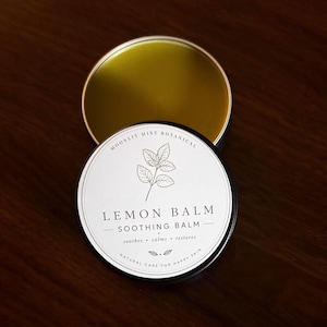 Può includere: Un barattolo di metallo rotondo e aperto di balsamo al limone con un'etichetta bianca. L'etichetta recita "LEMON BALM SOOTHING BALM" con le parole "soothes, calms, restores" sotto. Il barattolo è su una superficie marrone scuro.